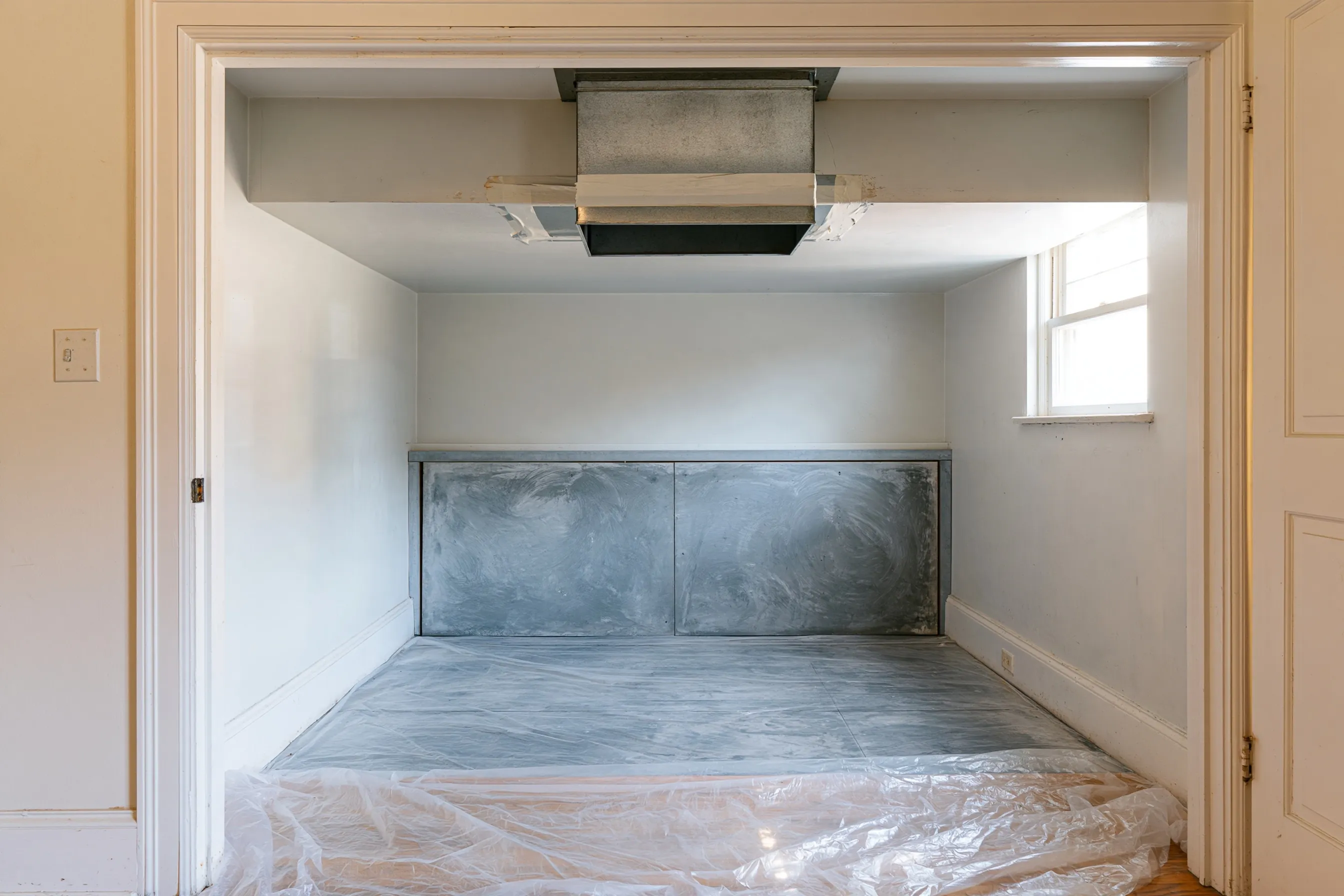 Empty furnace closet with a sheet-metal duct collar at the ceiling sealed with old white tape, a gray board panel on the back wall and plastic on the floor